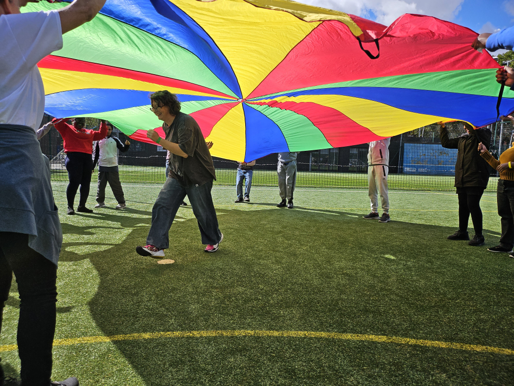 A person runs under a colourful parachute that other people are holding and swirling in the air.