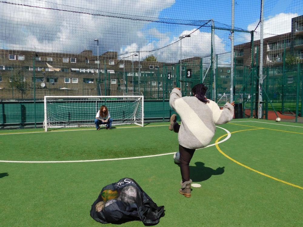 A lady kicks a football at the goal where there's another person trying to save it