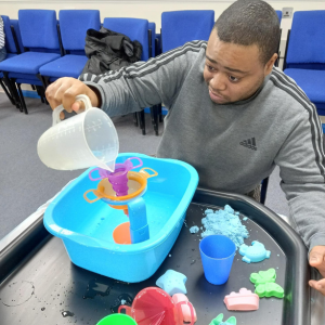 A man pours water into a bowl with plastic objects and foam, part of sensory play