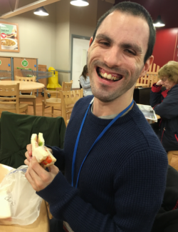 A man sits at a table laughing whilst he's enjoying a sandwich.