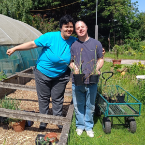 Two people stand in a garden, one holds two pots of plants