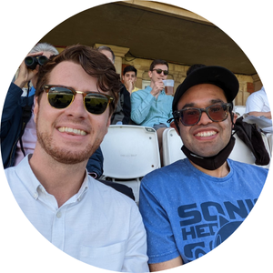 Two men sit in the stands watching a cricket game, both wearing sunglasses and smiling at the camera