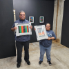 Two art students stand in a gallery space holding framed paintings