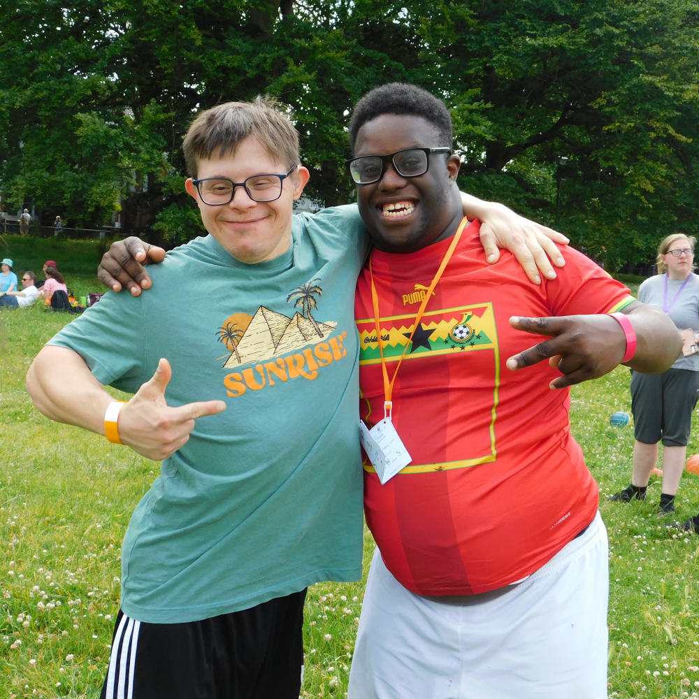 Two men stand with arms round each other's shoulders smiling at  the camera in a park