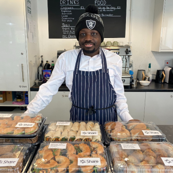 A man stands in a catering apron behind a table with a selection of food packaged up ready for an event Share is catering for