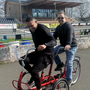 Two men ride on an adult sized tricycle in a velodrome