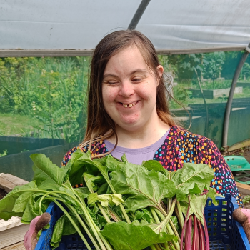 A lady stands in a polytunnel smiling and holding newly harvested vegetables