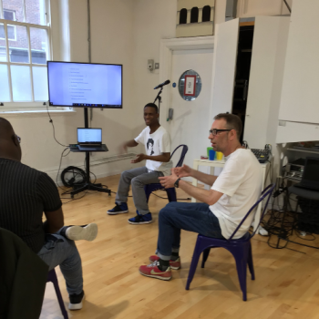 Three men sit in a circle with a screen behind them as they take part in a drama workshop