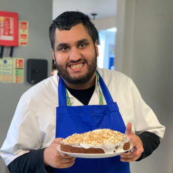 A man wearing an apron stands proudly holding a cake he's just baked