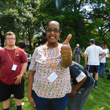 A lady stands in a park smiling at the camera and giving a thumbs up