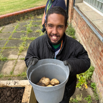 A man satnds outside with a bucket of potatoes he's just harvested.